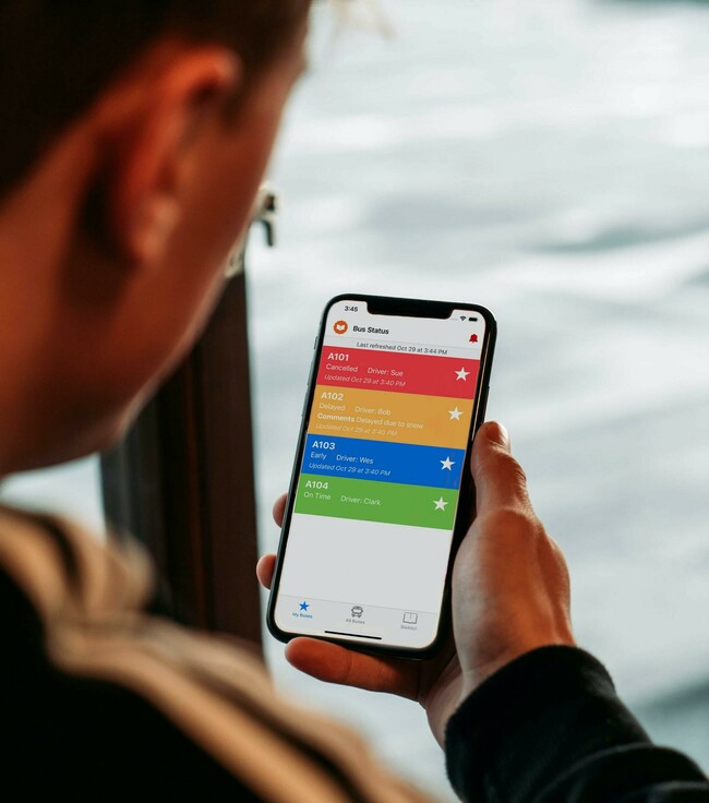 A man hold a phone. The phone screen shows the bus status listing, with buses cancelled, delayed, early, and on time.